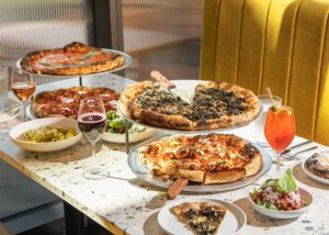Four pizzas with puffy, lightly charred crusts sit on a terrazzo table at a restaurant with cocktails, wine, appetizers, and a bit of a mustard-yellow banquette peeking through.