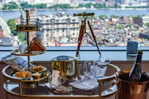 A gold-rimmed cart is topped with caviar and accoutrements, plus champagne, vodka, and glasses, in front of a big window with city skyline views.