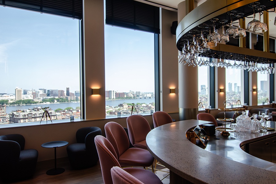 A curved bar has pink high-top seats, with large windows in the background looking out onto the Boston skyline.