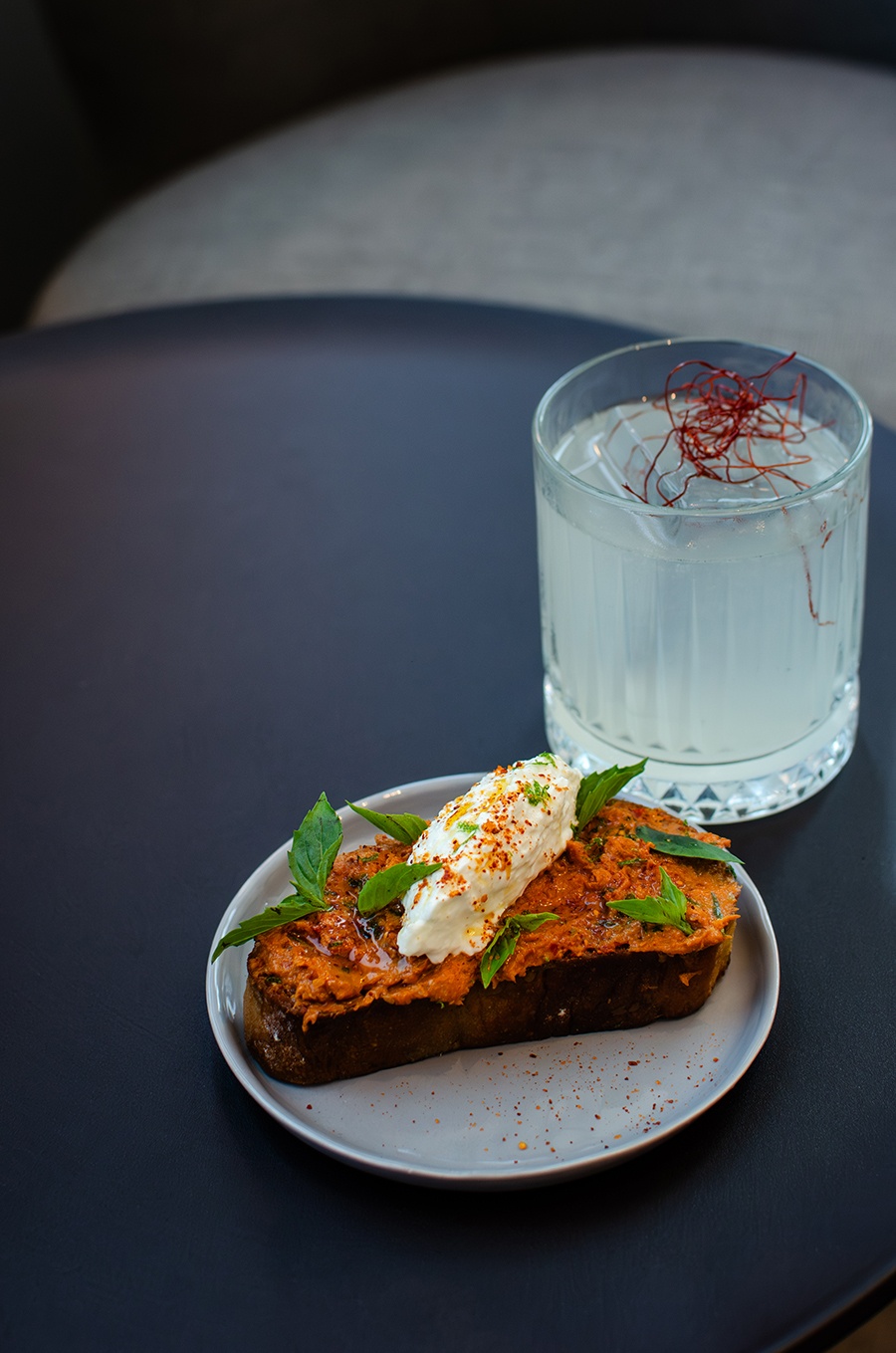 A piece of toast with a bright red spread and a quenelle of soft cheese sits next to a pale yellow-white cocktail garnished with saffron threads.