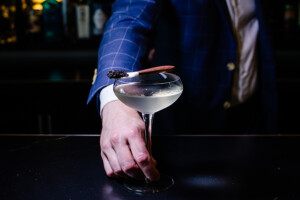 A bartender in a blue jacket holds the stem of a glass holding a pale white-yellow cocktail topped with a spoon full of caviar.